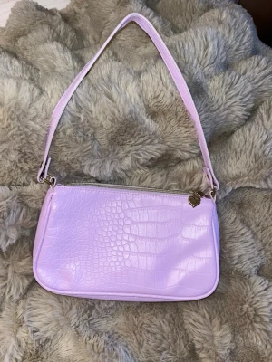 Ljuslila axelväska med krokopräglat mönster - Säljer en ljuslila axelväska i fejkläder med krokopräglat mönster. Väskan har en smal axelrem och dragkedja med ett gulligt hjärtformat hänge i guld. Perfekt för dig som vill ha en trendig och söt accessoar till din outfit.