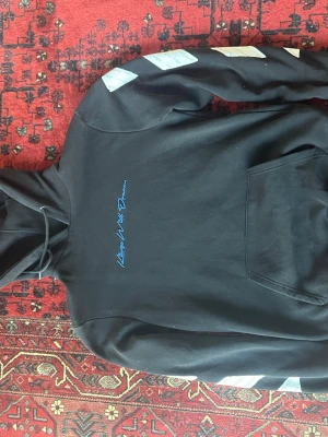 Kings Will Dream Hoodie Off-White med tryck - Svart hoodie från Kings Will Dream med blått broderat tryck 'Kings Will Dream' på bröstet och vita diagonala ränder på ärmarna. Klassisk känguruficka framtill och justerbar huva med snören. Tillverkad i mjuk bomull för skön känsla.