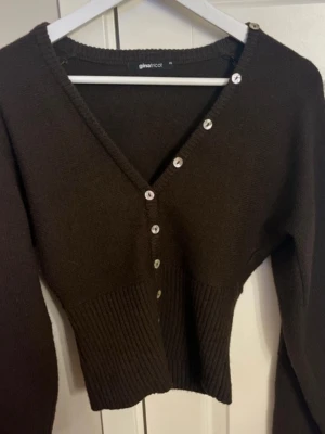 Brun kofta från Gina Tricot - Mörkbrun ribbstickad kofta från Gina Tricot med V-ringning och dekorativa vita knappar längs framsidan. Modellen har långa ärmar och bred ribbad kant nedtill för en snygg passform. Perfekt för lager på lager och enkel att styla. Helt ny 
