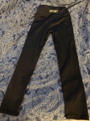 Svarta leggings med SPORT-tryck - Säljer ett par svarta leggings som är helt nya (aldrig använda )  med hög midja och SPORT-tryck baktill i vitt. De har en smal passform och är tillverkade i ett stretchigt material som sitter skönt på kroppen. Perfekta för träning eller chill dagar.