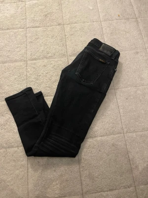 Svarta skinny jeans från nudie  - Säljer ett par svarta skinny jeans från nudie med klassisk femficksdesign och detaljerad söm vid knäna. Jeansen har en läderpatch bak i midjan och är tillverkade i stretchigt denimtyg för extra komfort.