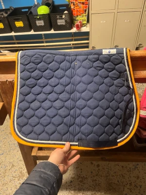 Marinblått schabrak med orange kant - Säljer ett marinblått schabrak med quiltat mönster och orange kantband. Schabraket har även en vit och grå rand längs kanten och kardborreband för enkel fastsättning. Perfekt till hästutrustningen och ger en snygg look i stallet.