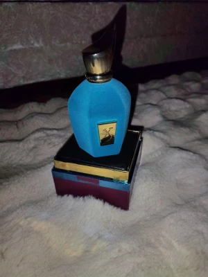 Xerjoff blå parfymflaska - Lyxig parfym från Xerjoff i en matt, blå flaska med gulddetaljer och elegant lock. Flaskan har en unik form och står på en exklusiv ask i lila och guld. Perfekt för dig som gillar exklusiva dofter och snygg design.