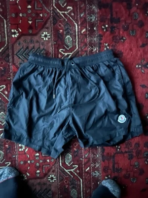 Svarta badbyxor från Moncler - Säljer ett par svarta badbyxor från Moncler. dragsko. Monclers logotyp sitter på vänster ben. Materialet är lätt och snabbtorkande, perfekt för stranden eller poolen. QR KOD OCH LABEL ÄR INKLUDERAT