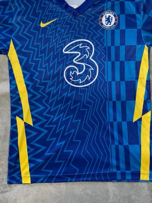 Chelsea FC matchtröja blå Nike M - Chelsea FC matchtröja i blått med gula detaljer och vitt tryck. Tröjan har klubbmärke på bröstet, Nike-logga och stort sponsortryck på framsidan. Tillverkad i lätt och ventilerande material, perfekt för fotboll eller träning.