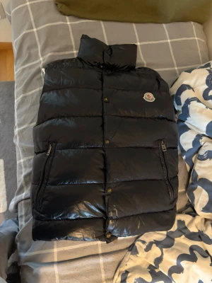 Svart dunväst från Moncler - Snygg svart dunväst från Moncler med hög krage och två dragkedjeförsedda fickor. Västen har ett quiltat mönster och Moncler-logga på bröstet. Perfekt för lager-på-lager och håller dig varm med sin fluffiga dunfyllning.