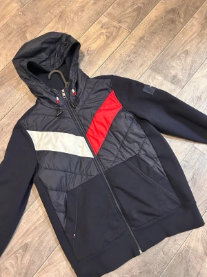 Marinblå cardigan - Snygg marinblå cardigan från Tommy Hilfiger med dragkedja, huva och justerbara snören. Framsidan har coola färgblock i rött och vitt. Köpt utomlands för ca 1000kr