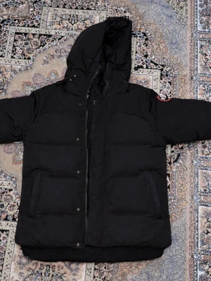 Svart dunjacka med huva Canada Goose - Säljer en svart dunjacka från Canada Goose med stor huva och två djupa fickor framtill. Jackan har en patch på ärmen och stängs med dragkedja och knappar. Perfekt för kalla vinterdagar och riktigt varm tack vare dunfyllningen.