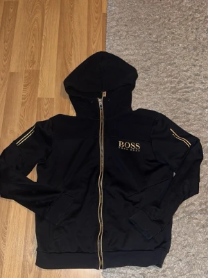 Svart hoodie från BOSS med gulddetaljer - Snygg svart hoodie från BOSS med dragkedja och guldfärgade detaljer. Loggan 'BOSS HUGO BOSS' syns på bröstet och det finns guldfärgade ränder på ärmarna. Hoodien har huva och fickor framtill. Perfekt för en stilren och modern look.