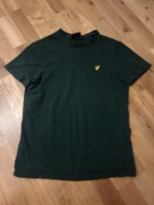 Svart t-shirt från Lyle & Scott - Klassisk svart t-shirt från Lyle & Scott med gul broderad logga på bröstet. T-shirten har rund hals och korta ärmar, perfekt basic-plagg till garderoben. Tillverkad i mjuk bomull för skön känsla.