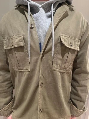 Snygg grön overshirt  - Stilren grön overshirt i kraftigt bomullstyg med två stora bröstfickor med lock och knapp. Klassisk krage och knappstängning framtill. Perfekt att bära över en hoodie för en avslappnad look. Jag är 186 cm lång och väger 75kg 