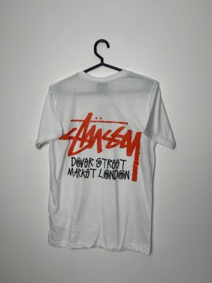 Vit Stüssy Dover Street Market tee - Säljer en clean vit t-shirt från Stüssy x Dover Street Market London. Tishan har klassisk passform, korta ärmar och tryck i rött och svart både fram och bak. Tillverkad i mjuk bomull, perfekt för dig som gillar streetwear och vill sticka ut.