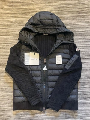 Svart Moncler cardigan - Säljer en snygg cardigan från moncler! Cardiganen är i mycket bra skick och har inga defekter förutom lite skavanker vid ena armen (se bild). Nfc-scan och Qr-kod funkar som den ska. Perfekt nu till vinter/vår väder! Om ni har några frågor så är de bara att höra av er😊