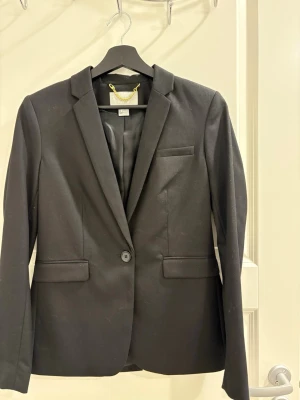 Svart klassisk kavaj med knapp - Stilren svart kavaj med en knapp framtill, smal passform och klassisk krage. Kavajen har två fickor med lock och en bröstficka. Fodrad insida och knappar vid ärmsluten. Perfekt för en clean och dressad look.