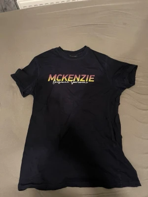 Svart McKenzie t-shirt med tryck - Snygg svart t-shirt från McKenzie med färgglatt tryck på bröstet där det står 'McKenzie Original Sportswear'. Klassisk passform och rund halsringning, perfekt till jeans eller shorts.