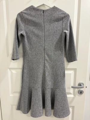 Grå klänning med volang från adL - Snygg gråmelerad klänning från adL med trekvartsärm och volang nedtill. Klänningen har en rak passform och dragkedja i ryggen. Perfekt för dig som gillar stilrena plagg med en twist.