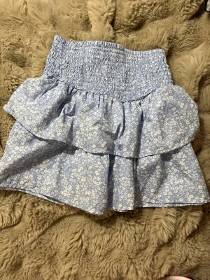 Blommig blå volangkjol från Gina Trickot  - Supersöt blå kjol från Gina med vita små blommor och bred smock i midjan. Kjolen har volangdetalj och är i ett lätt bomullstyg som passar perfekt till varmare dagar. Den har en kort modell och ger en fräsch och somrig vibe.