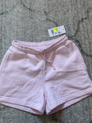 Ljusrosa shorts från Iconic Sportswear - Säljer ett par ljusrosa shorts från Iconic Sportswear med resårmidja och snörning. Mjukt material och avslappnad passform, perfekt för sommardagar. Tryck text 'Los Angeles' på benet för en extra detalj. Aldrig använda till och med har prislappen kvar. Original pris 120kr.