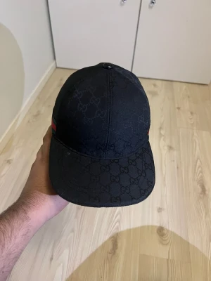 Svart Gucci keps med GG-mönster - Snygg svart keps från Gucci med det klassiska GG-monogrammet över hela kepsen. På sidan finns en grön och röd rand som ger en ikonisk touch. Böjd skärm och justerbar passform. Perfekt för dig som vill ha en stilren och lyxig accessoar.