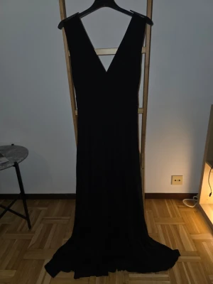 Svart långklänning med v-ringning - Elegant svart långklänning med djup v-ringning både fram och bak. Klänningen har ett flytande fall och är ärmlös, vilket ger en stilren och tidlös look. Perfekt för dig som vill ha en klassisk och enkel stil. Står tyvärr ingen storlek men passar mig som är S och är strechig i tyget så skulle kunna passa en M.