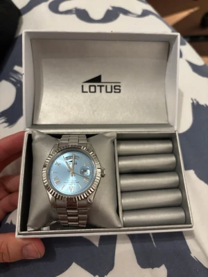 Silverfärgad klocka från Lotus - Stilren klocka från Lotus med silverfärgat metallarmband och ljusblå urtavla. Klockan har romerska siffror, dag- och datumvisning samt en räfflad bezel. Perfekt accessoar för dig som gillar klassisk men modern stil.