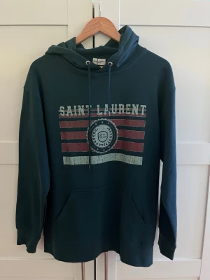 Mörkgrön Saint Laurent hoodie - Mörkgrön hoodie från Saint Laurent med tryck framtill i vitt och vinrött. 🛑Fråga gärna om du har funderingar såsom pris eller kräver fler bilder!