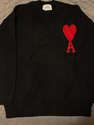 Svart stickad tröja från Ami Paris - Svart stickad tröja från Ami Paris med röd hjärt- och A-detalj på bröstet. Tröjan har rund halsringning, långa ärmar och ribbade muddar vid ärmslut och nederkant. Perfekt för dig som gillar stilrena plagg med en twist.