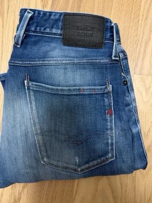 Blå jeans från Replay Denim - Säljer ett par blå jeans från Replay Denim med klassisk femficksmodell och snygga slitningar. Jeansen har en mörkblå tvätt, kontrastsömm sopch Replay-logga på bakfickan. Perfekta för en avslappnad och trendig look.
