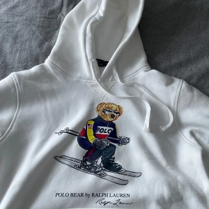 Vit hoodie Polo Bear Ralph Lauren - Vit hoodie från Ralph Lauren med den ikoniska Polo Bear-trycket där björnen åker skidor i färgglada kläder. Hoodien har huva med dragsko och en klassisk känguruficka framtill. Tillverkad i mjukt bomullsmaterial och har ribbade muddar vid ärmslut och nederkant. Storlek M men passar S