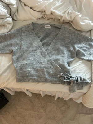 Grå omlottstickad tröja  - Säljer en ljusgrå stickad omlottopp från even&odd. Tröjan har långa ärmar, v-ringning och knytdetalj i sidan för en snygg passform. Perfekt att styla med jeans eller kjol för en chill look.