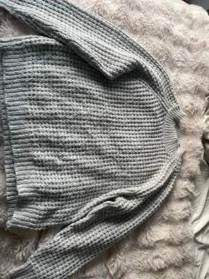 Ljusgrå stickad tröja med struktur - Mysig ljusgrå stickad tröja med grov struktur och rund hals. Tröjan har långa ärmar och ribbade muddar vid ärmslut och nederkant. Perfekt för lager på lager och enkel att matcha till jeans eller kjol.