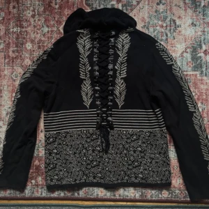 Svart kofta med snörning och beiga mönster. - Unik svart kofta med beiget mönster längs ärmar och nederkant, snörning baktill och volangkrage. Dragkedja framtill och dekorativa ränder över bröstet. 