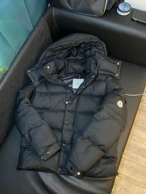 Svart Moncler dunjacka med huva - Säljer en svart dunjacka från Moncler med avtagbar huva och klassisk logotyp på ärmen. Jackan har tryckknappar, dragkedja och två fickor framtill. Fodret är glansigt och jackan har en sportig, puffig look som är riktigt trendig just nu.