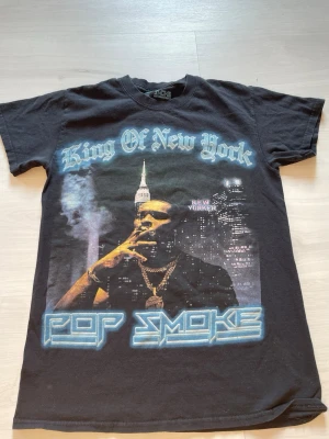 Pop Smoke King of New York T-shirt - Svart t-shirt med stort Pop Smoke-tryck på framsidan och texten 'King Of New York'. Baksidan har låtlista från albumet 'Meet The Woo'. Klassisk passform och rund hals. Perfekt för dig som gillar streetwear och hiphop.
