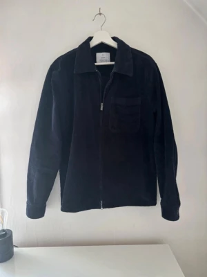 Blå manchester overshirt från bläck - Stilren blå overshirt i manchestertyg från bläck. Jackan har dragkedja framtill, klassisk krage och en bröstficka. Perfekt lager-på-lager-plagg med avslappnad passform och långa ärmar.