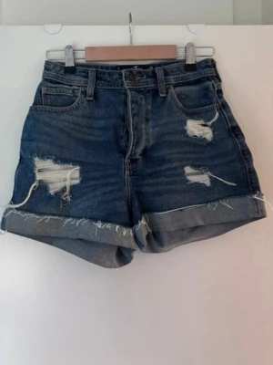 Blå denimshorts från Hollister - Snygga blå denimshorts med hög midja och råa, fransiga kanter. Shortsen har slitna detaljer framtill och uppvikta benslut för en avslappnad look. Klassisk femficksmodell med knapp och dragkedja.