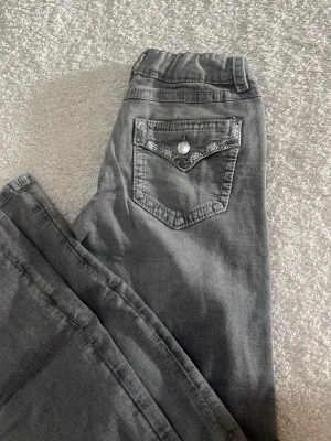 Grå bootcut jeans med spetsdetalj - Säljer ett par grå jeans med bootcut passform och snygg spetsdetalj på bakfickan. Jeansen har klassisk femficksdesign och knappstängning. Materialet är mjukt denim i bomull. Perfekta för dig som gillar en avslappnad men ändå trendig look.
