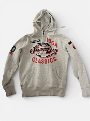 Grå Superdry hoodie med tryck - Snygg grå hoodie från Superdry med stort rött och svart tryck på bröstet där det står 'Superdry Classics 1954'. Hoodien har huva med dragsko, känguruficka och tryck även på ärmarna. Perfekt för en avslappnad streetwear-look.