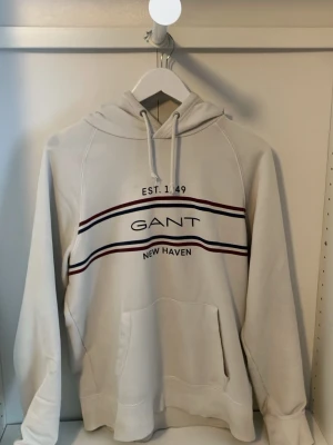 Vit hoodie från GANT med tryck - Säljer en vit hoodie från GANT med trycket 'EST. 1949 GANT NEW HAVEN' och två ränder i rött och blått över bröstet. Hoodien har huva med dragsko och en stor magficka. Tillverkad i mjukt bomullsmaterial, perfekt för en avslappnad stil.