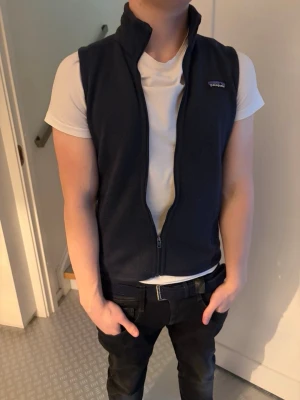 Mörkblå fleeceväst från Patagonia - Snygg mörkblå fleeceväst från Patagonia med hög krage och dragkedja framtill. Västen har två sidofickor och är tillverkad i mjukt fleecematerial. Perfekt att slänga över en t-shirt för en avslappnad look.