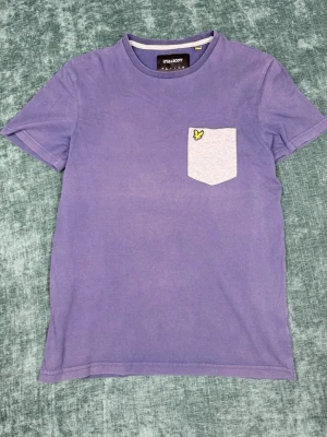 Lila t-shirt med ficka från Lyle & Scott - Snygg lila t-shirt från Lyle & Scott med kontrasterande ljusgrå bröstficka och den klassiska gula loggan broderad på fickan. T-shirten har rund halsringning och korta ärmar. Perfekt för en avslappnad och trendig stil.