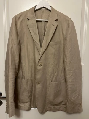 Beige kavaj från Riley - Beige Riley kavaj, strl 52. Ytter tyg - Linne 55%, Bomull 45% Foder - Polyester 100%. Ärmlängd (cm) 66 , Axelbredd (cm) 47.