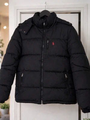 Svart pufferjacka från Ralph Lauren - Säljer en svart pufferjacka från Ralph Lauren med röd broderad logga på bröstet. Jackan har hög krage, dragkedja framtill och två fickor med dragkedja. Klassisk quiltad design och varm vaddering, perfekt för kalla dagar.