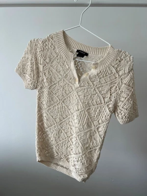 Beige stickad topp från Lindex  - Superfin beige stickad topp från Lindex  med korta ärmar och dekorativa knappar vid halsringningen. Snyggt mönstrad med ett luftigt, virkat utseende och ribbade kanter. Perfekt att styla med jeans eller kjol.