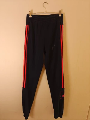 Adidas mjukisbyxor - Snygga svarta mjukisbyxor från Adidas med klassiska röda ränder längs benen och Adidas-logga på ena benet. Byxorna har resår i midjan och är tillverkade i mjukt material som passar perfekt för chill eller träning. Smal passform och sportig look. Storlek L för barn.