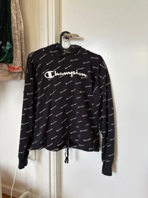 Svart hoodie från Champion - Snygg svart hoodie från Champion med vit logga framtill och all-over print av Champion-texten. Hoodien har huva med dragsko och långa ärmar med muddar. Perfekt för en avslappnad och sportig stil.