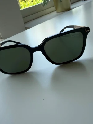 Svarta solglasögon från Tom Ford - Stilrena svarta solglasögon från Tom Ford med fyrkantig form och mörka glas. Bågarna är i svart plast med tunna guldfärgade metallskalmar som ger en lyxig känsla. Perfekta för att höja din look under soliga dagar.