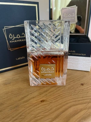Lattafa Khamrah Eau de Parfum 100ml - Lattafa Khamrah Eau de Parfum kommer i en elegant, fyrkantig glasflaska med ett unikt mönster och guldiga detaljer. Doften har en varm bärnstensfärg och flaskan levereras i en lyxig svart och guld presentförpackning. Perfekt för dig som gillar exklusiva parfymer.
