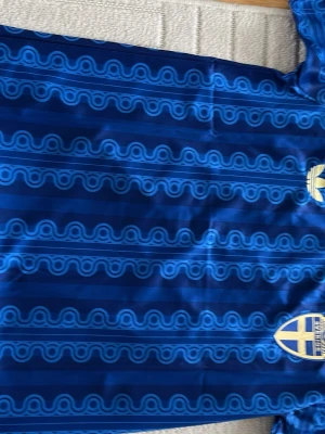 Blå Sverige fotbollströja Adidas - Säljer en blå fotbollströja från Adidas med Sveriges landslagsemblem på bröstet och mönstrade ränder i olika blå nyanser. Tröjan har korta ärmar och texten 'Sverige' i nacken. Perfekt för dig som vill visa ditt stöd för landslaget.