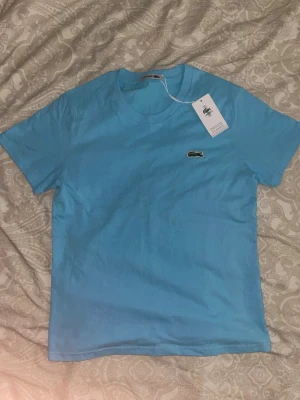 Blå t-shirt från Lacoste - Snygg ljusblå t-shirt från Lacoste med klassisk krokodillogga på bröstet. T-shirten har rund halsringning och korta ärmar, perfekt för en avslappnad stil. Tillverkad i mjuk bomull för maximal komfort.